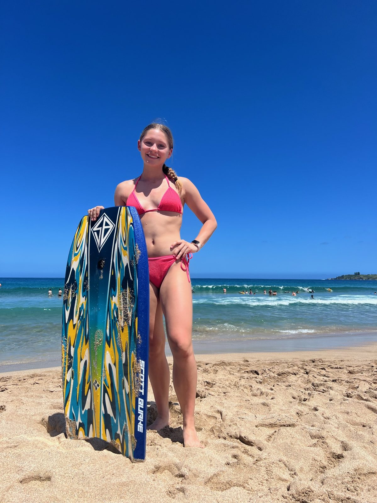 Teen with a boogie board on a sunny West Maui beach, swimmers and small surf behind